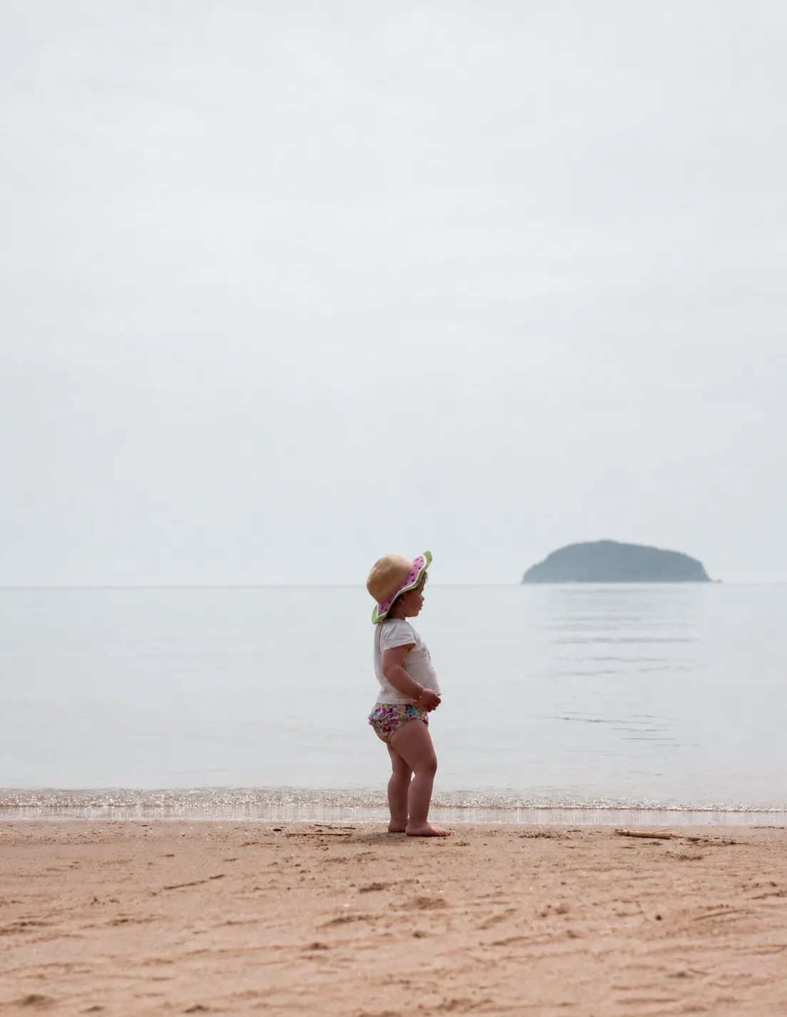 A small child standing at the edge of calm water on a wide, quiet beach