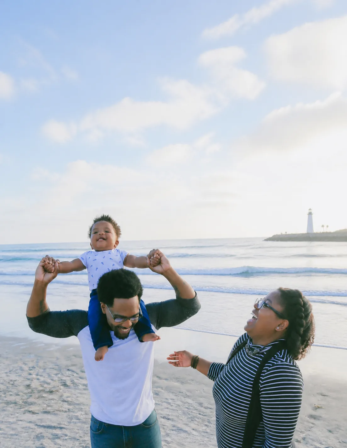 Two parents playing with a young child on the beach, a lighthouse behind them